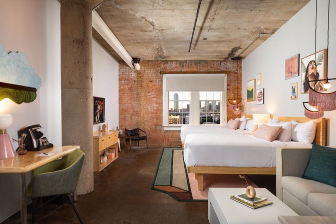 Sunlit industrial-chic loft hotel room with exposed brick and concrete ceiling, two queen beds with pastel pillows, eclectic wall art, desk, sofa and window framing a downtown urban skyline