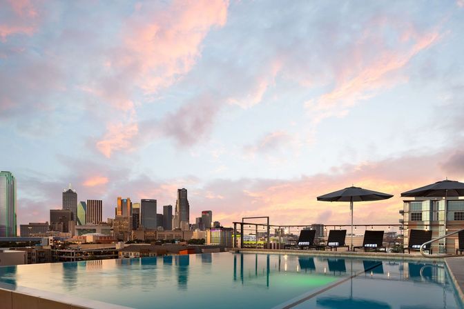 Rooftop infinity pool with lounge chairs and umbrellas overlooking a downtown skyline at a pastel pink sunset, city lights reflecting on the water.