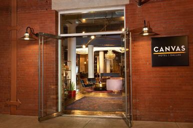 Open glass doors in a red-brick facade revealing a warm Dallas boutique hotel lobby with chandelier, velvet seating, white columns, and a patterned rug.