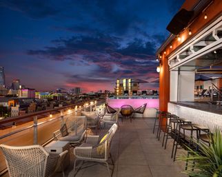 Rooftop bar patio overlooking a city skyline at twilight, with string lights, wicker lounge chairs, bar stools, and a colorful sunset sky.