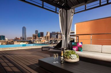 Rooftop pool terrace with cabana and modern lounge sofa, champagne flutes and succulent centerpiece on a coffee table, overlooking a downtown skyline under a clear blue sky.