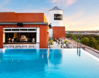 Sunset-soaked urban rooftop infinity pool with open-air bar, lounge seating and a white water tower overlooking treetops and city skyline.