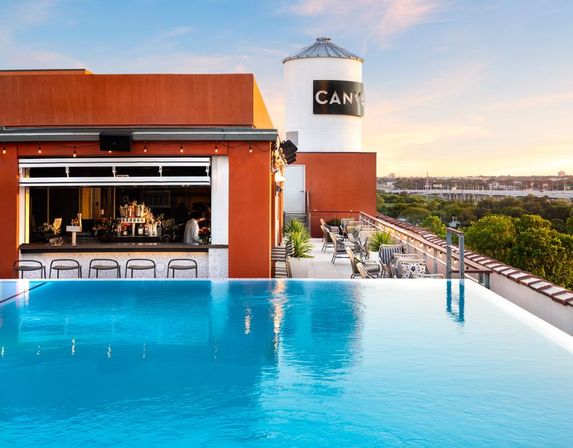 Sunset-soaked urban rooftop infinity pool with open-air bar, lounge seating and a white water tower overlooking treetops and city skyline.