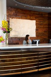 Smiling receptionist at a modern hotel lobby reception desk with curved wood-panel front, geometric wall art and tall orchid bouquet