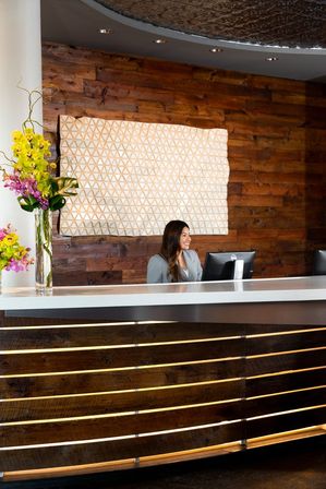 Smiling receptionist at a modern hotel lobby reception desk with curved wood-panel front, geometric wall art and tall orchid bouquet
