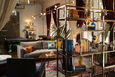 Cozy industrial urban loft living room with a gray sofa and colorful throw pillows, metal open-shelf room divider filled with books and vases, patterned area rug, deep red velvet curtains, pool table in the background and warm ambient lighting.