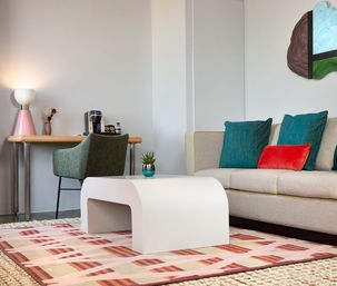 Stylish modern apartment living room with beige sofa, teal and red throw pillows, white curved coffee table on a patterned rug, green desk chair at a wooden workspace with lamp and coffee maker, and a small potted plant.