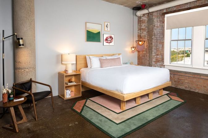 Cozy industrial loft bedroom with exposed brick and concrete column, large window with city view, wooden platform bed with crisp white linens, nightstand and lamp, geometric green-and-pink area rug, modern wall art and hanging pendant lights.