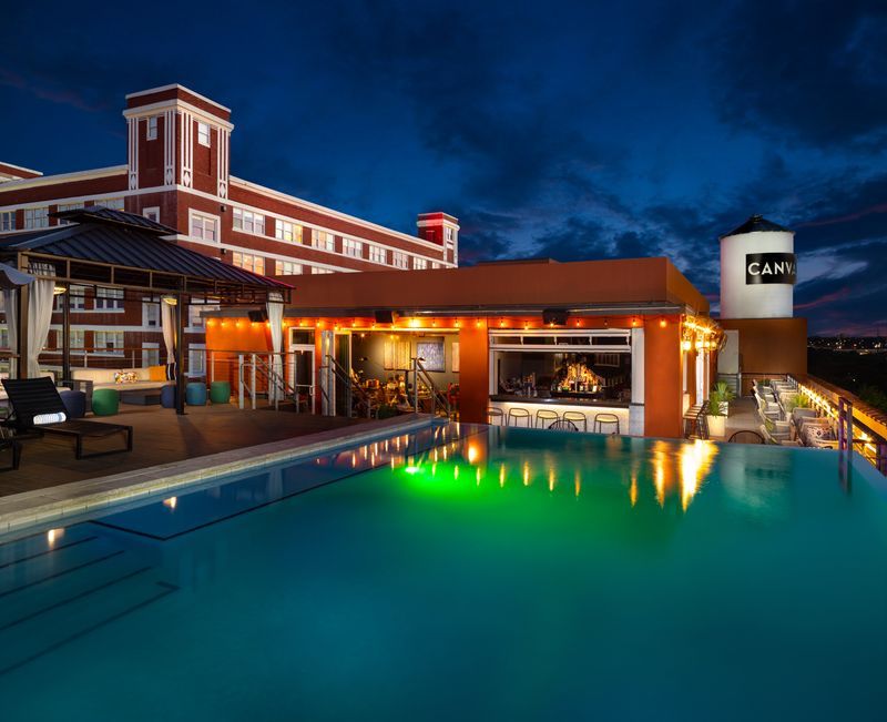 Glowing rooftop infinity pool and lively rooftop bar on a hotel terrace at dusk, with illuminated seating, historic brick building and deep‑blue city skyline.