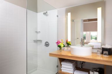 Bright, spa-like modern bathroom with glass walk-in shower, white tiled walls, wooden vanity with round vessel sink, illuminated mirror and a vase of colorful flowers.