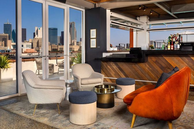Rooftop lounge with modern seating — orange velvet sofa, patterned armchairs and round ottomans — a wood-accent bar and floor-to-ceiling windows framing a downtown city skyline and outdoor patio.