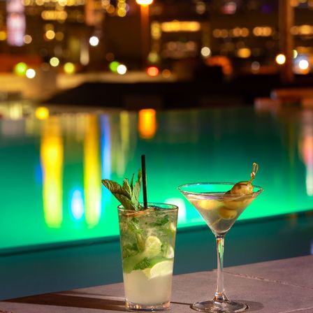 Two cocktails by a glowing rooftop pool at night with blurred city skyline lights — a mint mojito with lime and straw and a martini garnished with olives, vibrant bokeh reflections.