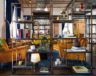 Open metal shelving filled with books, glass vases and decor framing a modern industrial loft lounge with mustard velvet armchairs, blue curtains, and a patterned blue rug