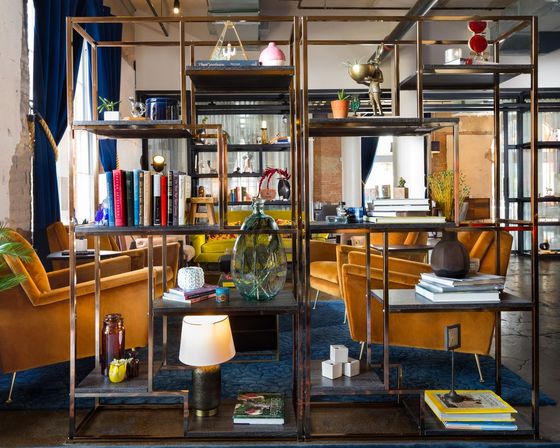 Open metal shelving filled with books, glass vases and decor framing a modern industrial loft lounge with mustard velvet armchairs, blue curtains, and a patterned blue rug