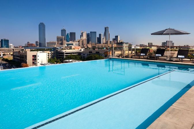 Rooftop infinity pool with bright turquoise water, chaise lounges and umbrella on the deck, overlooking a sunlit downtown city skyline under a clear blue sky.