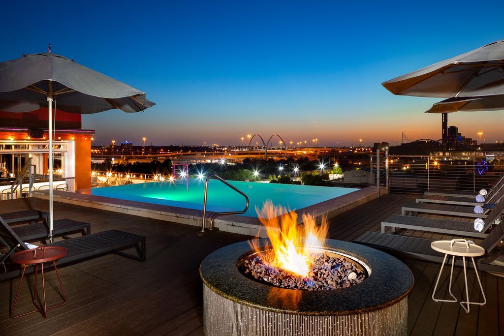 Rooftop infinity pool glowing turquoise at dusk with a roaring fire pit, lounge chairs and umbrellas overlooking an illuminated city skyline and arched bridge.