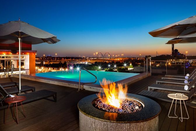 Rooftop infinity pool glowing turquoise at dusk with a roaring fire pit, lounge chairs and umbrellas overlooking an illuminated city skyline and arched bridge.