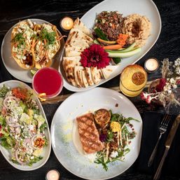 Overhead restaurant spread with seared salmon on greens and grilled lemon, veggie slaw tacos, pita and hummus platter with quinoa and crudités, mixed salad, colorful cocktails, floral garnish and lit candles on a dark table.