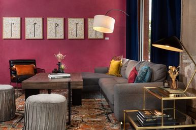Stylish city loft living room with a magenta accent wall, gray sectional sofa with colorful pillows, wooden coffee table, patterned area rug, framed artwork, velvet ottomans and brass side table with lamps.