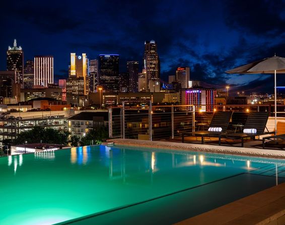 Rooftop infinity pool glowing teal at night with lounge chairs and umbrella on the deck, downtown skyline lights reflecting in the water under a deep blue sky.
