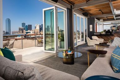 Sunny rooftop lounge with folding glass doors, modern sofas and coffee tables opening onto an outdoor terrace with chairs and a downtown city skyline under a clear blue sky.