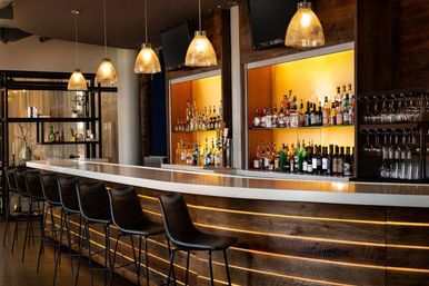 Sleek modern bar interior with long white countertop, row of black leather bar stools, backlit wood shelving stocked with liquor bottles and wine, amber pendant lights and hanging glassware.