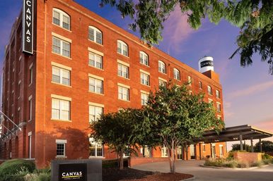 Red-brick historic hotel building with tree-lined covered entrance and rooftop water tower glowing at sunset in Dallas, Texas