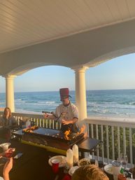 Beachfront terrace teppanyaki chef in a red hat performing a flaming grill show on a balcony overlooking ocean waves and a sandy beach, diners watching during golden hour.
