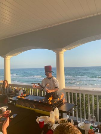 Beachfront terrace teppanyaki chef in a red hat performing a flaming grill show on a balcony overlooking ocean waves and a sandy beach, diners watching during golden hour.