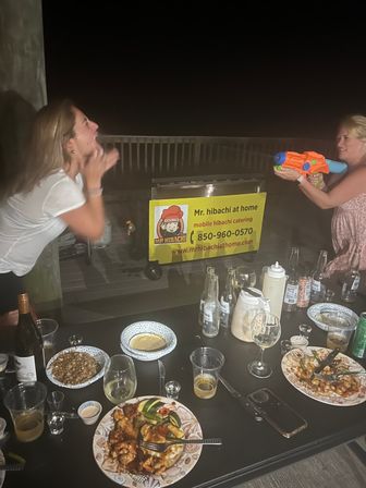 Two women laughing on a dark outdoor deck at night; one sprays a colorful water gun toward a portable hibachi cart, while a table in the foreground is strewn with plates of grilled food, drinks and bottles.