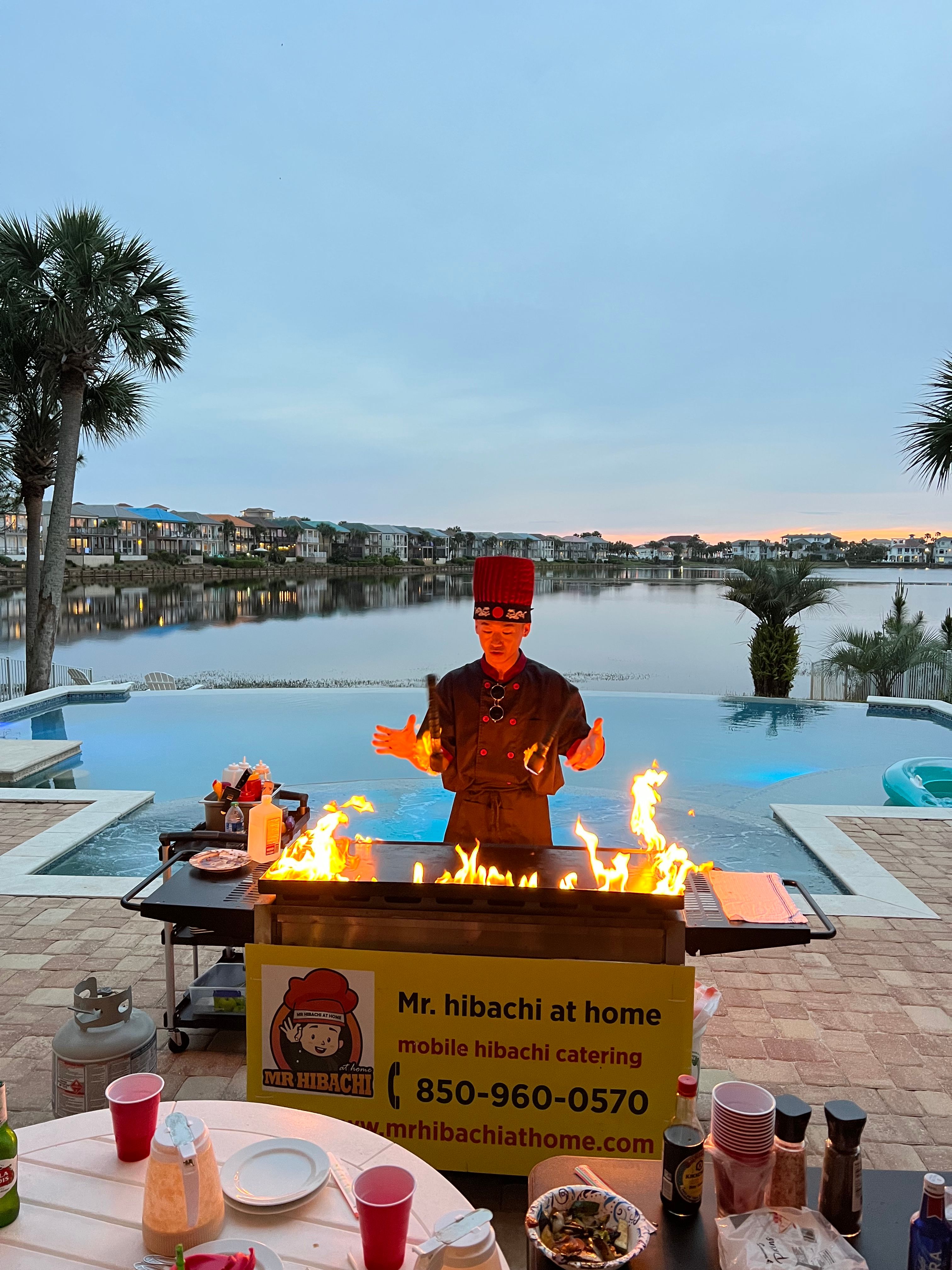 Poolside hibachi chef in a red hat cooking over a flaming griddle by a calm waterfront at sunset, palm trees and lakeside homes in the background.