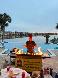 Poolside hibachi chef in a red hat cooking over a flaming griddle by a calm waterfront at sunset, palm trees and lakeside homes in the background.