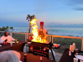 Beachfront hibachi chef in a red hat creating a towering flame on a teppanyaki grill on a seaside deck at sunset, diners at tables, a palm tree and calm ocean waves in the background.