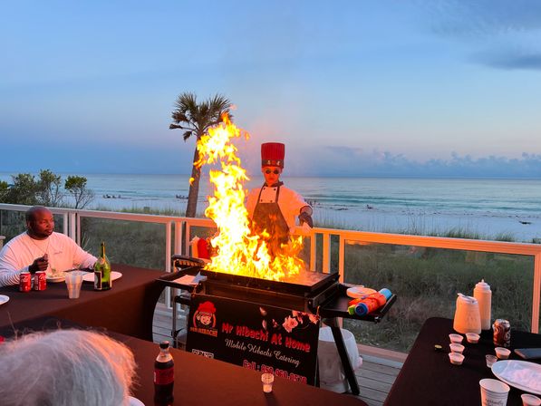 Beachfront hibachi chef in a red hat creating a towering flame on a teppanyaki grill on a seaside deck at sunset, diners at tables, a palm tree and calm ocean waves in the background.