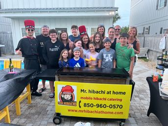 Smiling group of adults and kids at an outdoor patio celebration gathered around a mobile hibachi grill with a bright yellow catering sign; two chefs in tall red hats ready to cook.