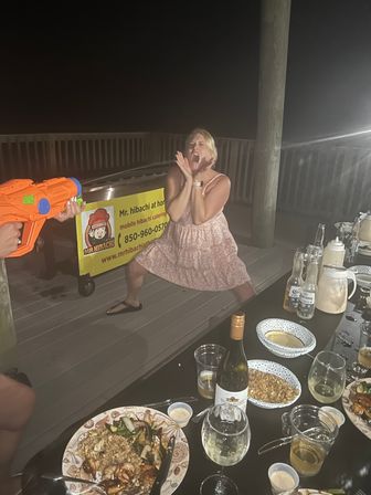 Nighttime outdoor deck party: woman in a pink sundress playfully screaming as an orange water gun is aimed at her beside a hibachi-style cart, with plates of fried rice, skewers, wine and beer on a long table.