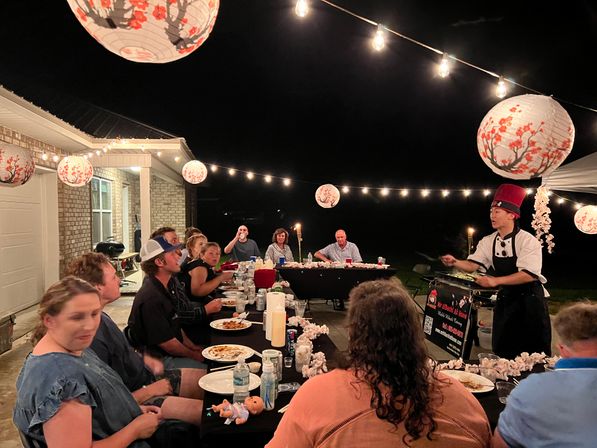 Backyard outdoor dinner party at night: guests seated at a long table under string lights and floral paper lanterns while a hibachi-style chef in a red hat cooks on a griddle.