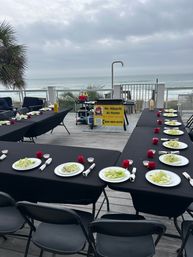 Oceanside deck hibachi setup with long black-clothed tables, plated salads and rolled napkins, a portable grill station and cloudy beach view