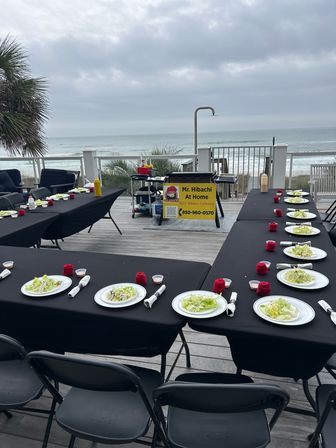Oceanside deck hibachi setup with long black-clothed tables, plated salads and rolled napkins, a portable grill station and cloudy beach view
