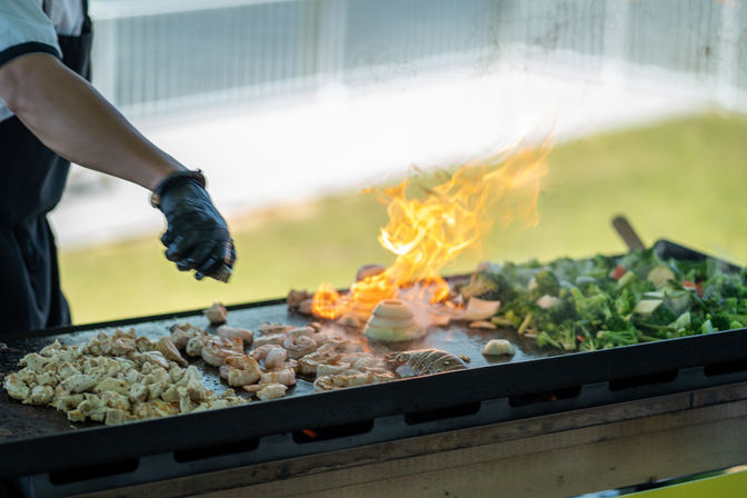 Gloved cook seasoning shrimp, diced chicken and mixed vegetables on a flaming outdoor flat-top griddle at a food stall