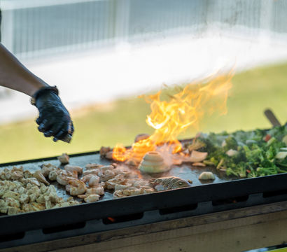 Gloved cook seasoning shrimp, diced chicken and mixed vegetables on a flaming outdoor flat-top griddle at a food stall