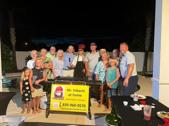 Smiling multigenerational group poses poolside at night around a hibachi chef cooking on a griddle at a backyard patio party.
