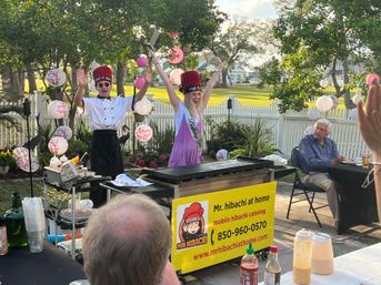 Backyard hibachi party with two chefs in tall red hats performing at a griddle while a birthday guest wearing a sash cheers amid pink paper lanterns and seated guests by a white picket fence.