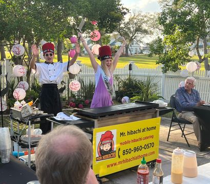 Backyard hibachi party with two chefs in tall red hats performing at a griddle while a birthday guest wearing a sash cheers amid pink paper lanterns and seated guests by a white picket fence.
