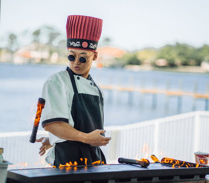 Hibachi chef in a tall red hat and round sunglasses theatrically flips a flaming spatula over a teppanyaki grill at a sunny waterfront food stall.