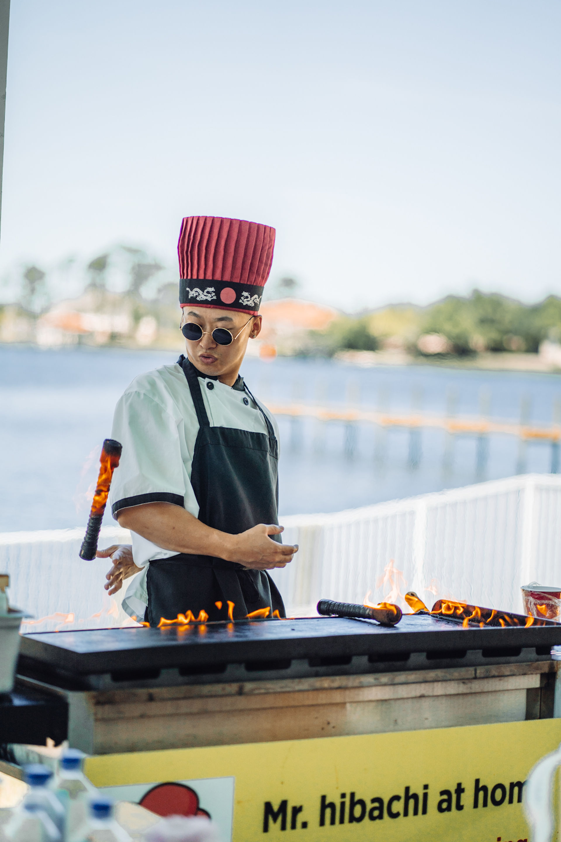 Hibachi chef in a tall red hat and round sunglasses theatrically flips a flaming spatula over a teppanyaki grill at a sunny waterfront food stall.