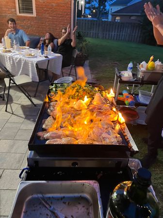 Backyard patio barbecue at dusk with a flaming flat-top griddle cooking chicken and vegetables while guests sit at a table outdoors