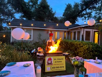 Backyard hibachi dinner at dusk with a chef in a red hat cooking over a dramatic flame on a griddle, string lights and paper lanterns overhead, set tables and floral centerpieces in front of a house