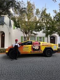 Chef in a red hat and black apron posing beside a bright orange-and-yellow wrapped electric pickup with a cartoon chef logo, parked on a cobblestone residential driveway in front of a white house with green shutters.