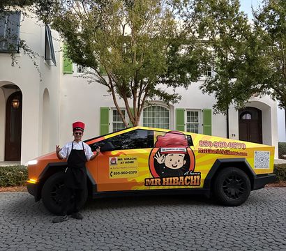 Chef in a red hat and black apron posing beside a bright orange-and-yellow wrapped electric pickup with a cartoon chef logo, parked on a cobblestone residential driveway in front of a white house with green shutters.